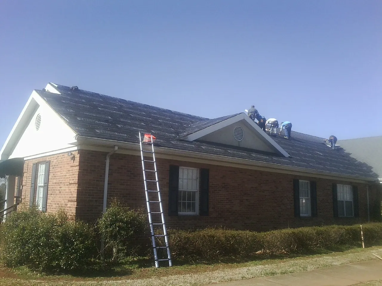 Crew tearing shingles off a brick ranch roof with a ladder set — Emergency Roof Repair work by our crew in Chalmette