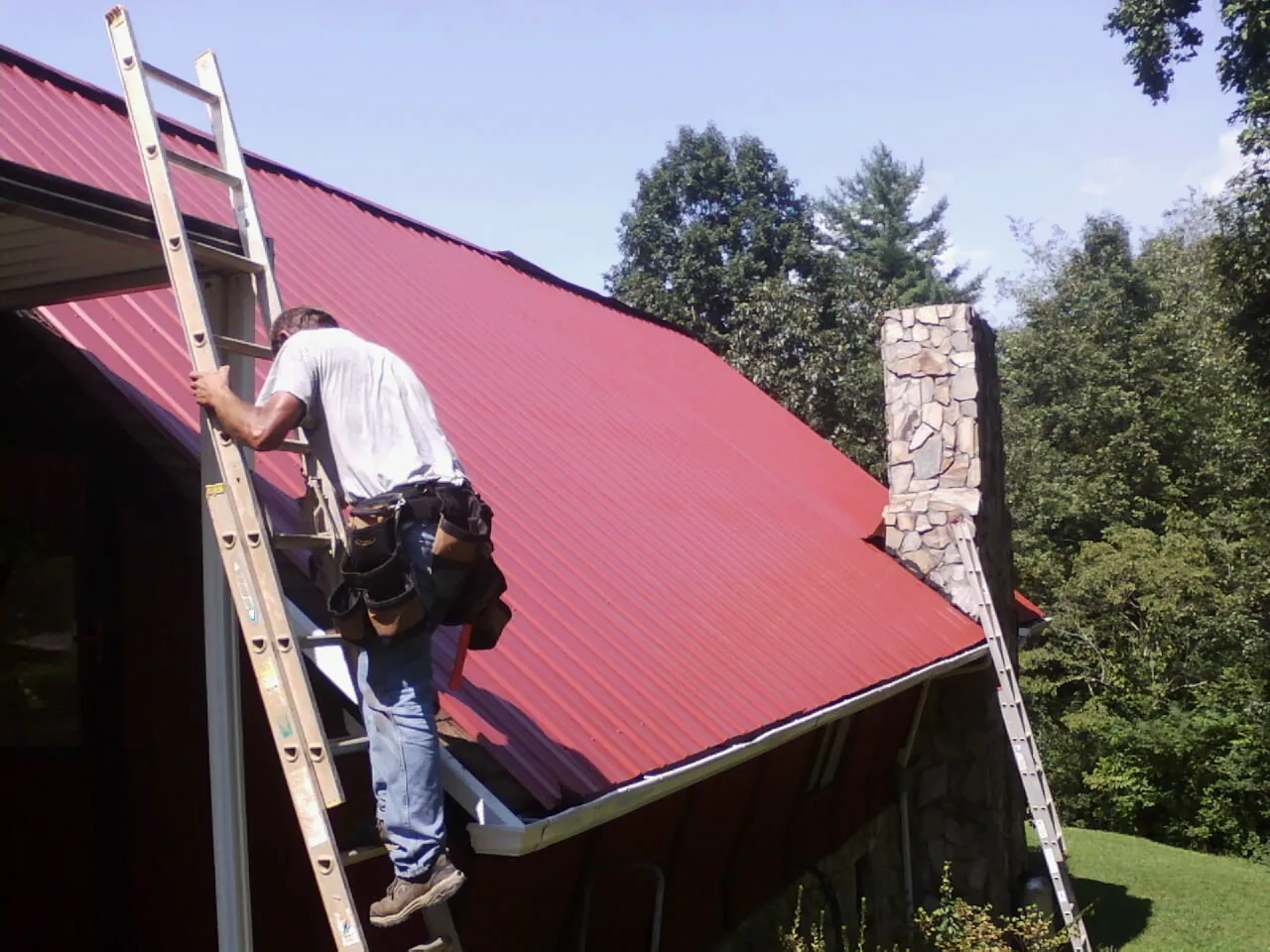 Roofer on a ladder beside a red metal roof and stone chimney — Soffit & Fascia Repair by Roofing Brothers in Chalmette